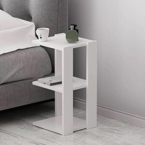 May include: A white bedside table with two shelves and a square base. A mug, a book, and a green bottle are on the table. The table is next to a gray bed.