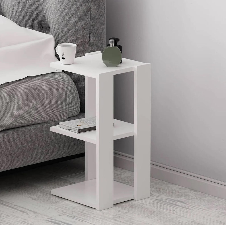 May include: A white bedside table with two shelves and a square base. A mug, a book, and a green bottle are on the table. The table is next to a gray bed.