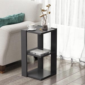 May include: A dark gray, modern side table with two shelves. A white teacup and saucer sit on the top shelf, along with a clear vase holding cotton stems. A book rests on the lower shelf. The table is next to a light-colored sofa.