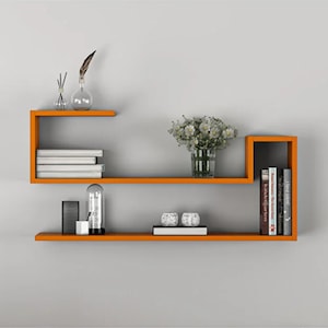 May include: A bright orange wall-mounted shelving unit with two levels. The top shelf holds a stack of white books and a clear glass vase with a feather and white flowers. The bottom shelf holds a black rectangular object, a clear glass dome, two white decorative spheres, and a row of books.