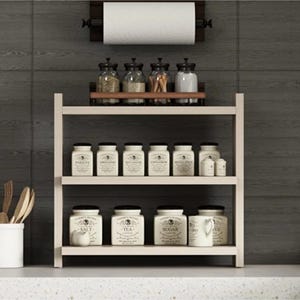 May include: A light-colored wooden spice rack with three tiers, holding various labeled ceramic jars and glass containers. A paper towel holder is mounted above the rack. The jars feature black text labels and lids, organized neatly on the shelves.