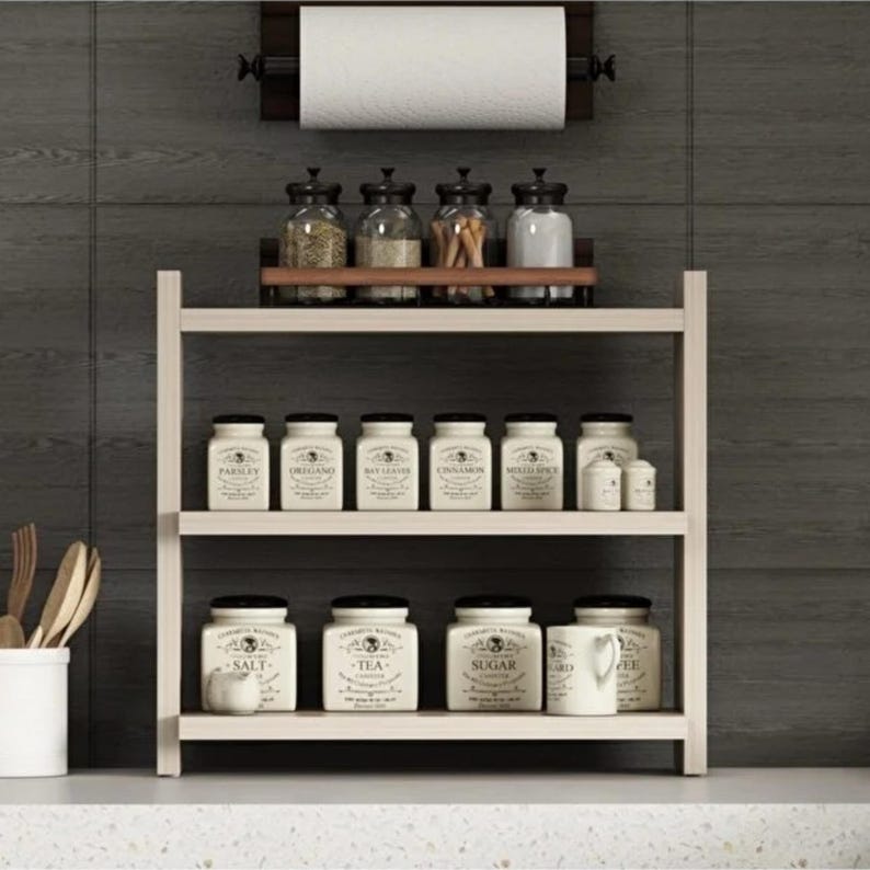 May include: A light-colored wooden spice rack with three tiers, holding various labeled ceramic jars and glass containers. A paper towel holder is mounted above the rack. The jars feature black text labels and lids, organized neatly on the shelves.