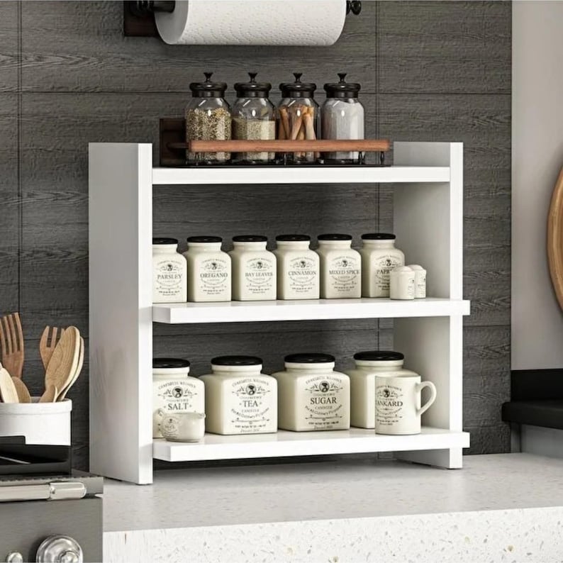 May include: A white two-tiered wooden spice rack holding various glass and ceramic jars. The top shelf features four glass jars with black lids. The middle and bottom shelves contain ceramic jars labeled with names like Salt, Tea, and Sugar.