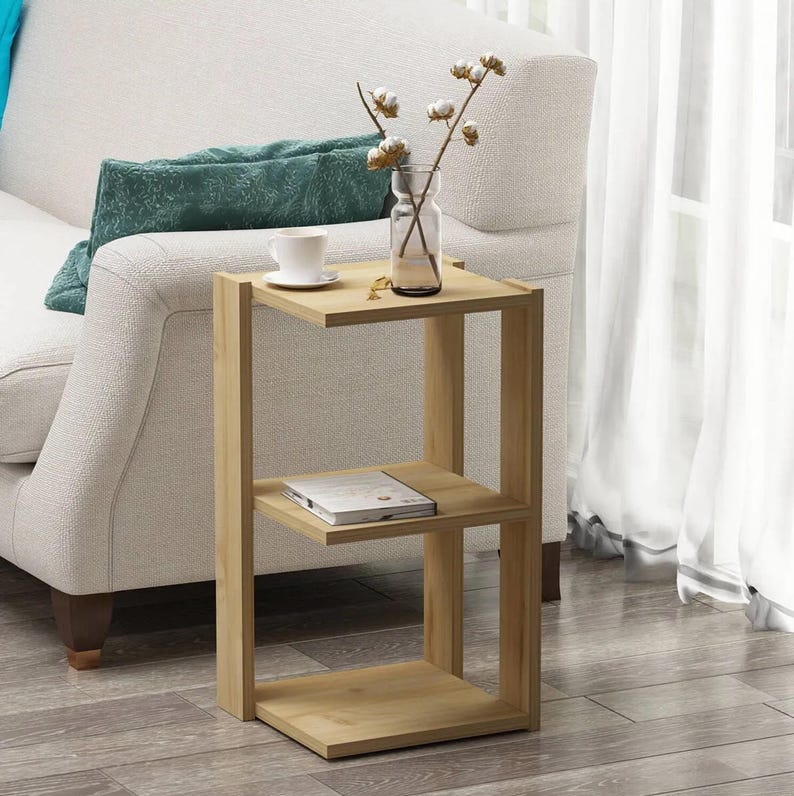 May include: A light-colored wooden side table with three shelves. A small glass vase with cotton stems, a teacup, and a book are on the shelves. The table is next to a white sofa with a teal pillow.