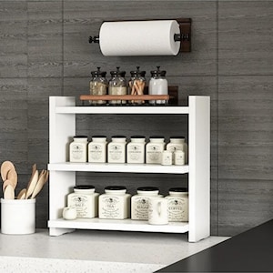 May include: A white two-tiered wooden spice rack holding twelve labeled ceramic jars, with a wall-mounted paper towel holder above. A white utensil crock containing wooden spoons sits to the left of the rack against a dark gray tiled wall.