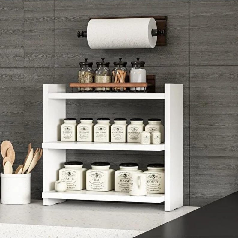 May include: A white two-tiered wooden spice rack holding twelve labeled ceramic jars, with a wall-mounted paper towel holder above. A white utensil crock containing wooden spoons sits to the left of the rack against a dark gray tiled wall.