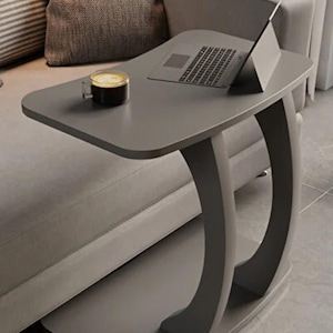 May include: A grey side table with a curved base and a rectangular top. A laptop and a cup of coffee are on the table. The table is next to a grey sofa and a patterned cushion.