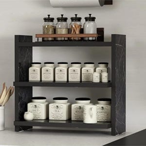 May include: A three-tiered black marble-patterned spice rack holding four glass jars with black lids on the top shelf, and eight cream-colored ceramic canisters labeled with various spices and pantry staples on the middle and bottom shelves.