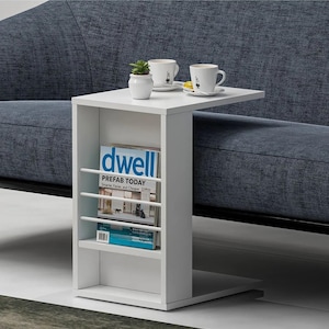 Op de afbeelding: Een witte bijzettafel met een tijdschriftenrek en een tafelblad. Twee witte koffiekopjes en een kleine plant in een pot staan op het tafelblad. Een tijdschrift met de titel "dwell" staat in het rek. De tafel staat naast een blauwe bank.