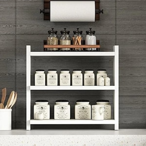 May include: A white three-tiered wooden shelf holding twelve white ceramic spice jars with black labels and a small white pitcher. Above the shelf, a wall-mounted wooden paper towel holder supports a roll of white paper towels.