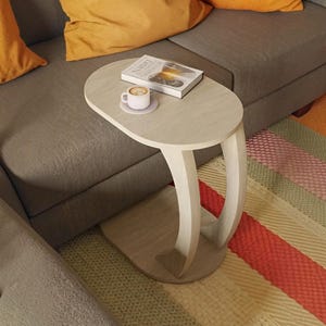 May include: A light-coloured wood side table with an oval top and a curved, C-shaped base. The table holds a white ceramic mug on a matching saucer and a book with the title Running Dawn visible on the cover, resting on the smooth tabletop surface.