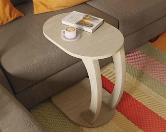 May include: A light-colored wood side table with an oval top and a curved, C-shaped base. The table holds a white ceramic mug on a matching saucer and a book with the title Running Dawn visible on the cover, resting on the smooth tabletop surface.