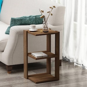 Puede incluir: Una mesa auxiliar de madera con diseño en forma de C. La mesa tiene tres niveles, con una taza y un jarrón con flores en el estante superior. Un libro descansa sobre el estante del medio. La mesa está junto a un sofá blanco.