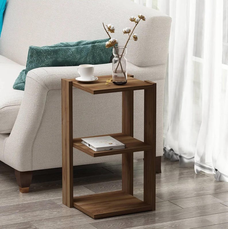 May include: A wooden side table with a C-shaped design. The table has three levels, with a cup and a vase with flowers on the top shelf. A book rests on the middle shelf. The table is next to a white sofa.