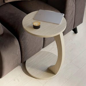 Può includere: Un tavolino a forma di C color legno chiaro con piano e base circolari. Sulla superficie sono appoggiati un tablet argentato e una piccola tazza di vetro contenente una bevanda scura con schiuma. Un supporto verticale curvo collega i due livelli.