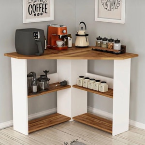 Op de afbeelding: Een hoekkast met een lichtbruin houten blad en witte steunen. De kast heeft planken en bevat een koffiezetapparaat, een airfryer, een waterkoker en kruidenpotjes. De muur erboven heeft ingelijste kunst.