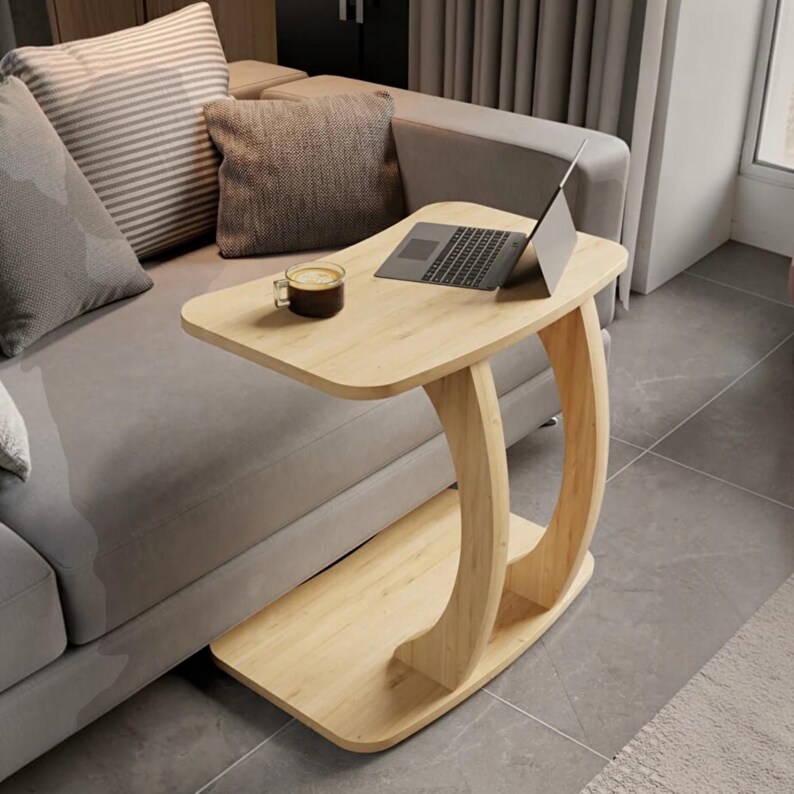 May include: A light-colored wooden C-shaped side table with a smooth rectangular top and a curved support base. A dark gray laptop is open on the surface next to a clear glass mug containing a dark brown beverage with a light foam top.