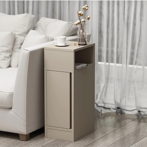 May include: A beige side table with a small cabinet and open shelf. A white teacup and saucer sit on top, next to a glass vase with cotton stems. The table is positioned next to a white sofa.