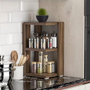 May include: A two-tier wooden corner shelf unit featuring a dark brown finish. The top shelf holds five glass spice jars with silver lids, while the bottom shelf contains four glass spice jars with wooden lids. A small green plant sits on top.