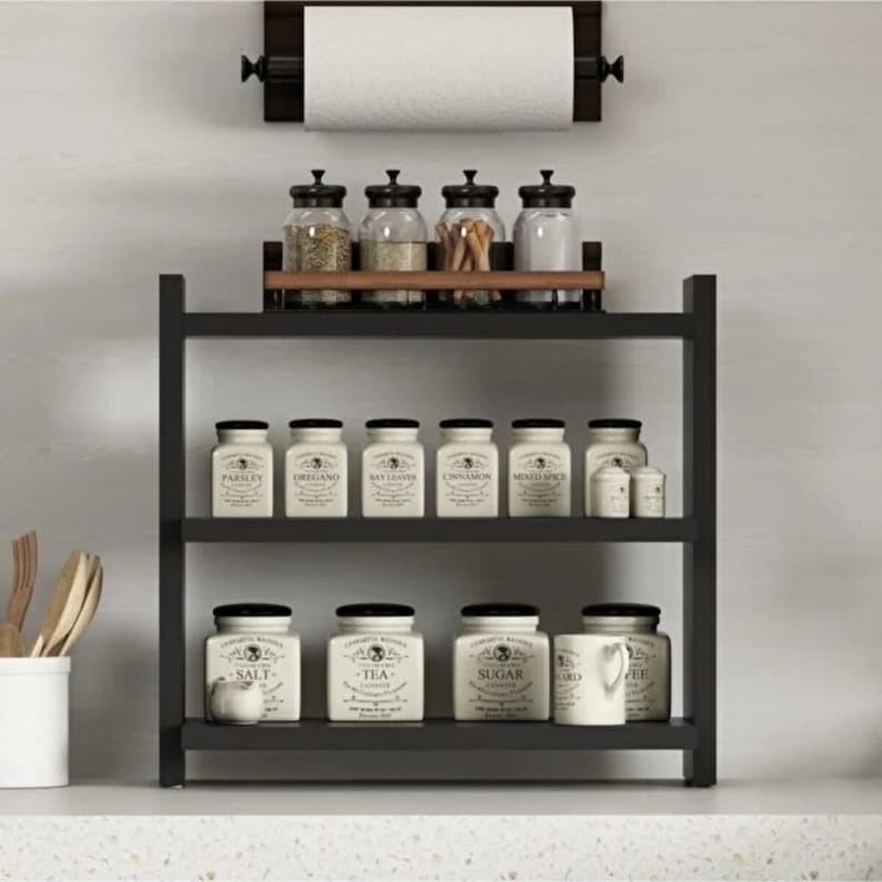 May include: A black three-tiered kitchen shelf holding white ceramic canisters labeled with various spices and ingredients, topped with four glass jars and a wall-mounted paper towel holder with a roll of white paper towels.