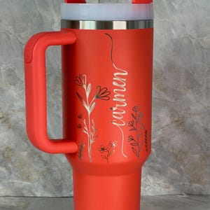 May include: A red insulated travel mug with a handle, a white lid, and a straw. The mug features the name "Carmen" in cursive and floral patterns. The mug is designed for cold drinks.