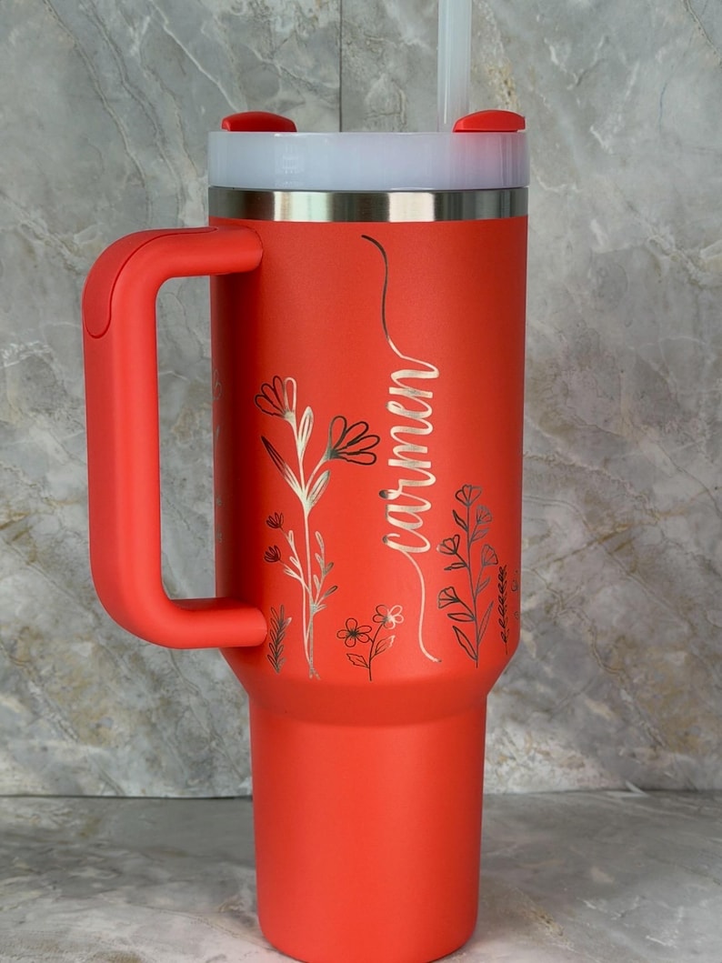 May include: A red insulated travel mug with a handle, a white lid, and a straw. The mug features the name "Carmen" in cursive and floral patterns. The mug is designed for cold drinks.