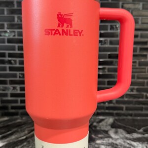 May include: A red Stanley Quencher H2.0 tumbler with a handle and straw. The tumbler has a white and silver lid and a white label with black text. The tumbler is 1.18 litres.