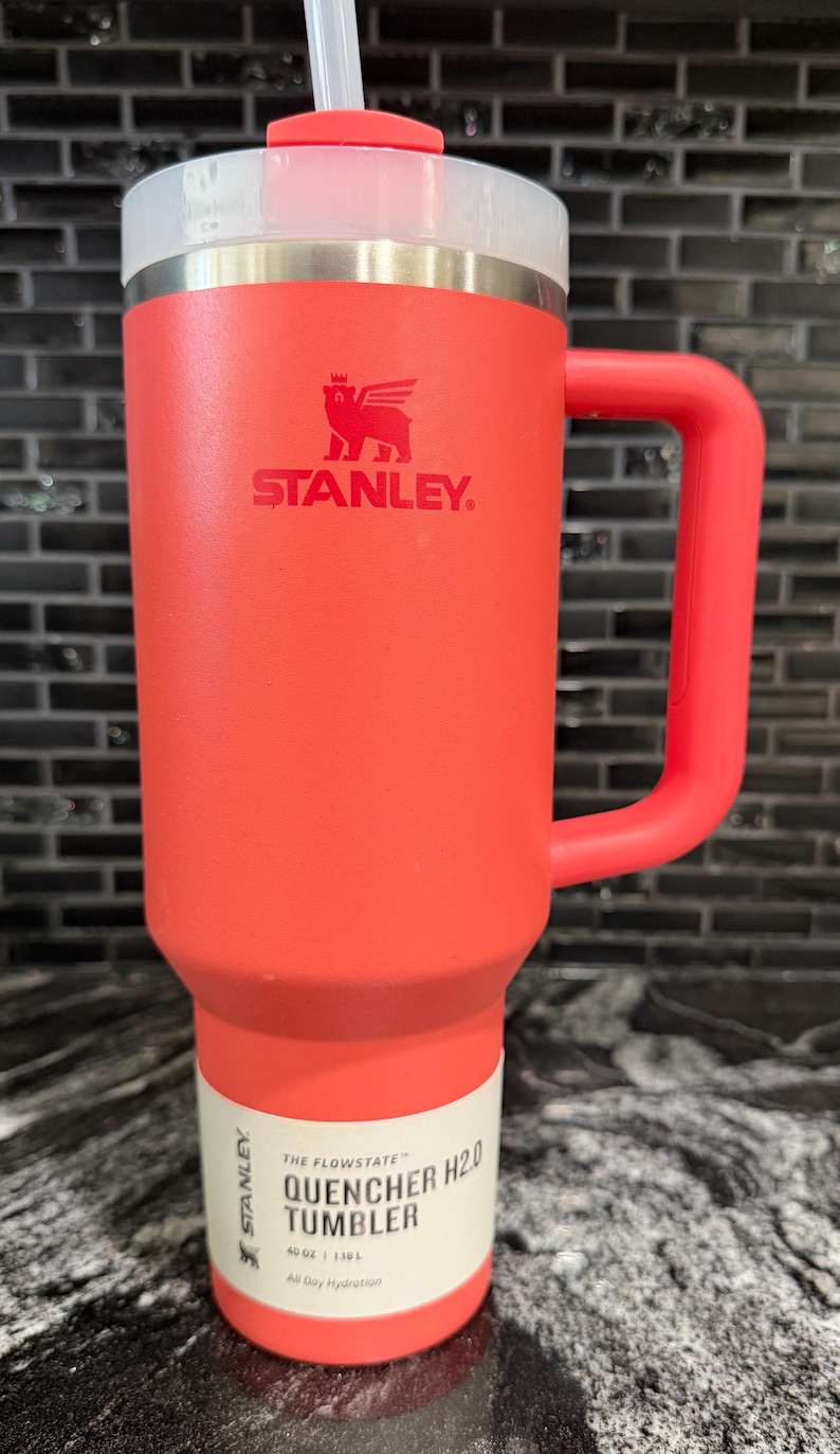 May include: A red Stanley Quencher H2.0 tumbler with a handle and straw. The tumbler has a white and silver lid and a white label with black text. The tumbler is 1.18 litres.