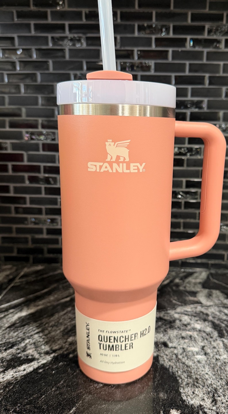 May include: A coral-coloured Stanley Quencher H2.0 Flowstate tumbler with a handle and a white lid. The tumbler has a straw and the Stanley logo. The tumbler is 1.18L and is designed for all-day hydration.