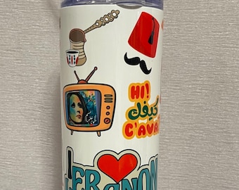 May include: White tumbler with a variety of colorful stickers. The stickers include a red fez hat, a black mustache, and the word "Lebanon" with a red heart. A metal straw extends from the lid.