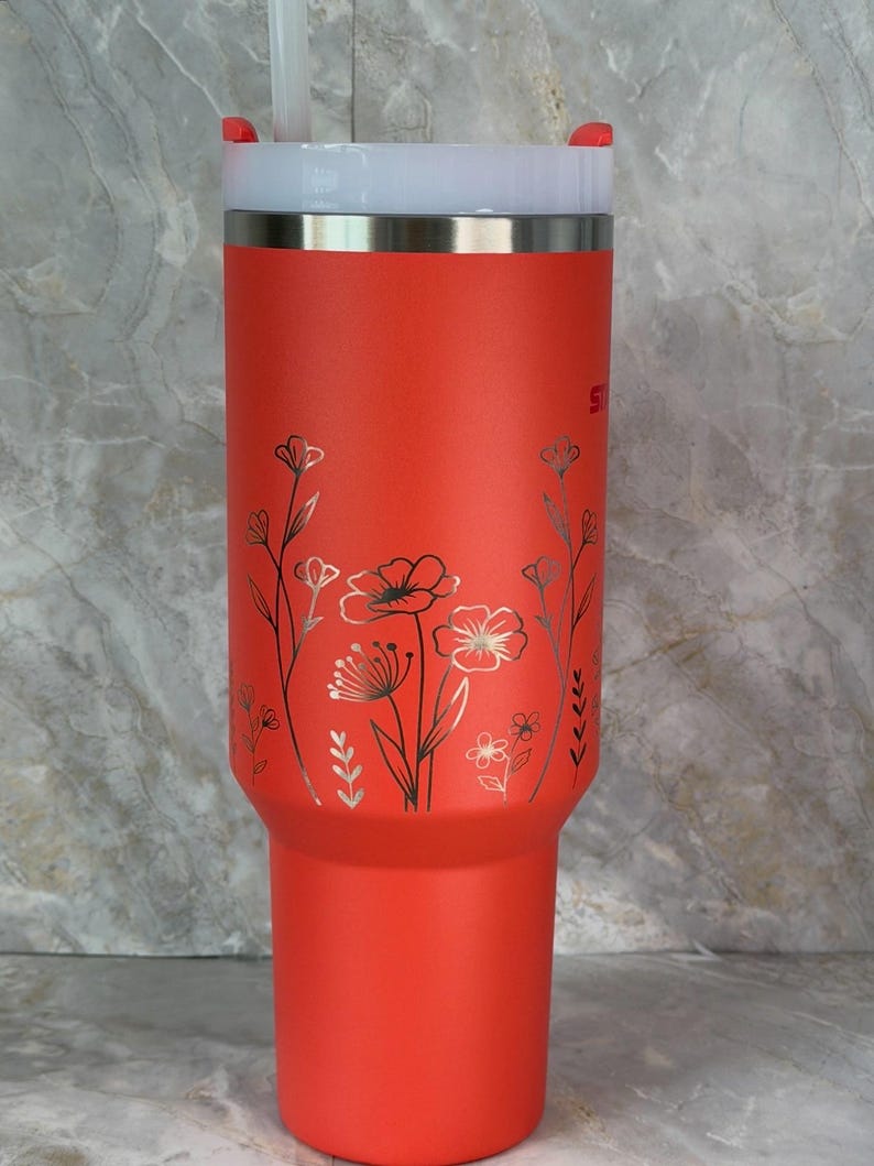 May include: A bright red insulated tumbler with a white lid and a silver rim. The tumbler has a floral design in silver and black. A white straw is inserted into the lid. The tumbler is designed to keep drinks cold or hot.