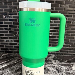 May include: A green Stanley Quencher H2.0 tumbler with a white lid and handle. The tumbler has a straw and the Stanley logo. The label reads "Quencher H2.0 Tumbler" and indicates a 1.18L capacity.