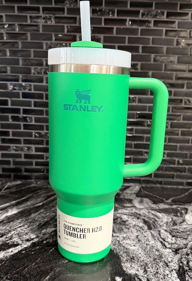 May include: A green Stanley Quencher H2.0 tumbler with a white lid and handle. The tumbler has a straw and the Stanley logo. The label reads "Quencher H2.0 Tumbler" and indicates a 1.18L capacity.