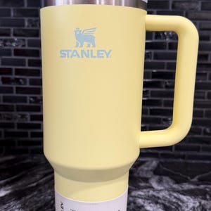 May include: A yellow Stanley Quencher H2.0 tumbler with a handle and straw. The tumbler has a white lid and a white label with the text "The Flowstate Quencher H2.0 Tumbler". The tumbler is 1.18L.