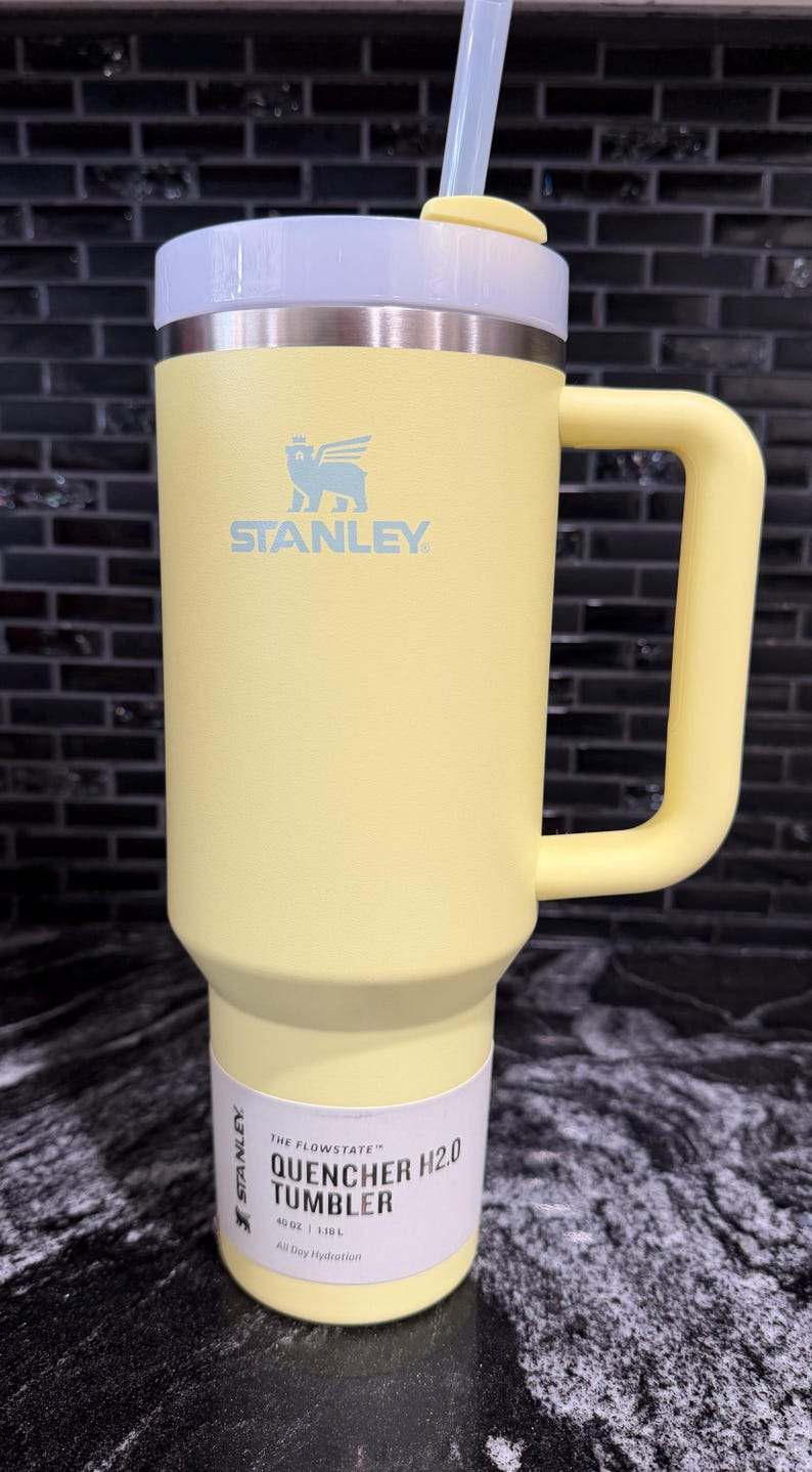 May include: A yellow Stanley Quencher H2.0 tumbler with a handle and straw. The tumbler has a white lid and a white label with the text "The Flowstate Quencher H2.0 Tumbler". The tumbler is 1.18L.