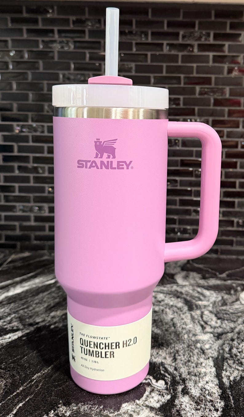 May include: A pink Stanley Quencher H2.0 Tumbler with a handle, lid, and straw. The tumbler features the Stanley logo and the text "THE FLOWSTATE QUENCHER H2.0 TUMBLER" on the label. It holds 1.18 litres.