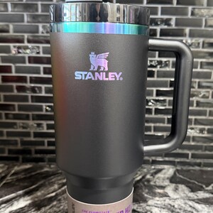 May include: Black Stanley Quencher H2.0 Flowstate tumbler with a handle and straw. The tumbler has a black body with a holographic iridescent band around the top. The Stanley logo is visible on the front. The tumbler holds 1.18L.