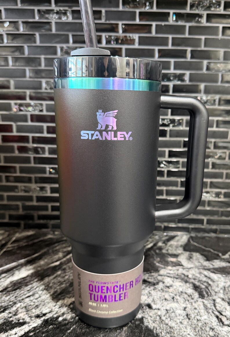 May include: Black Stanley Quencher H2.0 Flowstate tumbler with a handle and straw. The tumbler has a black body with a holographic iridescent band around the top. The Stanley logo is visible on the front. The tumbler holds 1.18L.