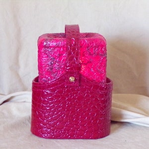 May include: A pink faux crocodile skin makeup bag with a gold handle and a matching pink box inside.
