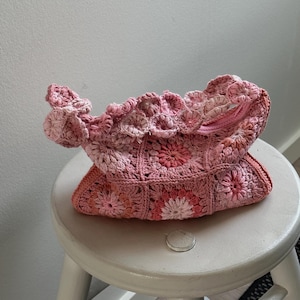 以下が含まれることがあります： 花柄のピンクの編み物バッグ。バッグは白いスツールの上に置かれています。