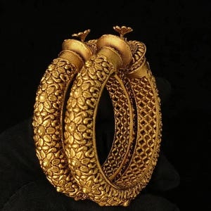 Peut inclure: Deux bracelets rigides dorés présentant un motif floral texturé sur la surface extérieure et un design en treillis sur la bande intérieure. Chaque bracelet est surmonté d'un petit fleuron décoratif en forme de fleur au point de fermeture.