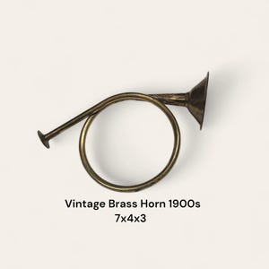 May include: Vintage brass horn from the 1900s. The horn is a warm brass color with a circular body and a flared bell. The text on the image reads "Vintage Brass Horn 1900s 7x4x3".