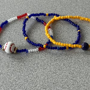 May include: Three beaded bracelets featuring small round glass beads in red, white, blue, and yellow. One bracelet includes a white spherical bead with red stitching resembling a baseball, while another features a single dark blue round bead.