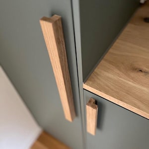 Könnte beinhalten: Nahaufnahme von zwei Holztürgriffen, einer vertikal und einer horizontal, montiert an einer Salbeigrünen Schranktür. Die Türgriffe sind aus hellem Holz gefertigt und haben ein schlichtes, rechteckiges Design.