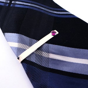 May include: A silver tie clip with a pink gemstone. The tie clip is on a striped tie with black, white and blue stripes.