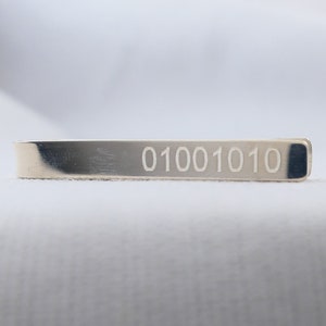 Pode incluir: Um prendedor de gravata de prata com o código binário "01001010" gravado nele.