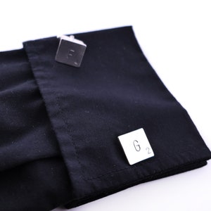May include: Two silver cufflinks with engraved letters. The cufflinks are on a black shirt cuff. The cufflink on the left has the letter "F" engraved on it. The cufflink on the right has the letters "G 2" engraved on it.
