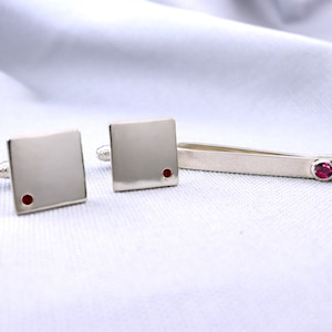 May include: A set of silver cufflinks and a tie bar, each featuring a small red gemstone.