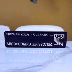May include: A rectangular black plaque featuring white text that reads BRITISH BROADCASTING CORPORATION and MICROCOMPUTER SYSTEM. To the right of the text is a white graphic of an owl composed of small dots, reflecting on the surface below.
