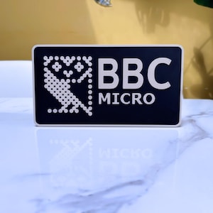 May include: A rectangular black sign with a white border featuring a white pixelated owl graphic on the left side and the white text BBC MICRO on the right side. The sign has a smooth finish and is displayed in a horizontal orientation.
